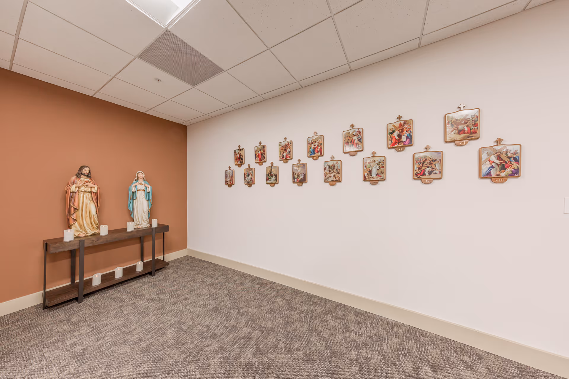 A quiet room with religious decor including statues of Jesus and Mary on a wooden table with candles, and a series of framed religious images mounted on a white wall. The room has a brown accent wall and carpeted floor.