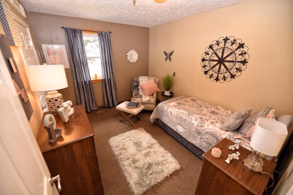A cozy bedroom with a single bed covered in a patterned comforter and multiple pillows. There is a wooden dresser with a lamp and decorative items on it, a small wooden nightstand with a lamp and decor, a cushioned chair with a pink pillow and a blanket, and a small ottoman. The room has beige walls with decorative wall hangings including a large circular metal piece and a butterfly. A window with blue curtains lets in natural light, and a fluffy white rug lies on the carpeted floor.