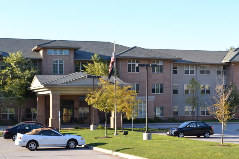 Exterior view of a three-story assisted living residence building with a covered entrance, several windows, and a few small trees in front. There are three cars parked in the parking lot near the entrance, and an American flag is flying on a flagpole near the center of the image.