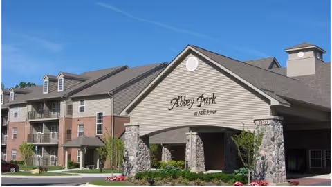 Front entrance of a multi-story senior living building with a covered porte-cochère and a sign reading "Abbey Park at Mill River".