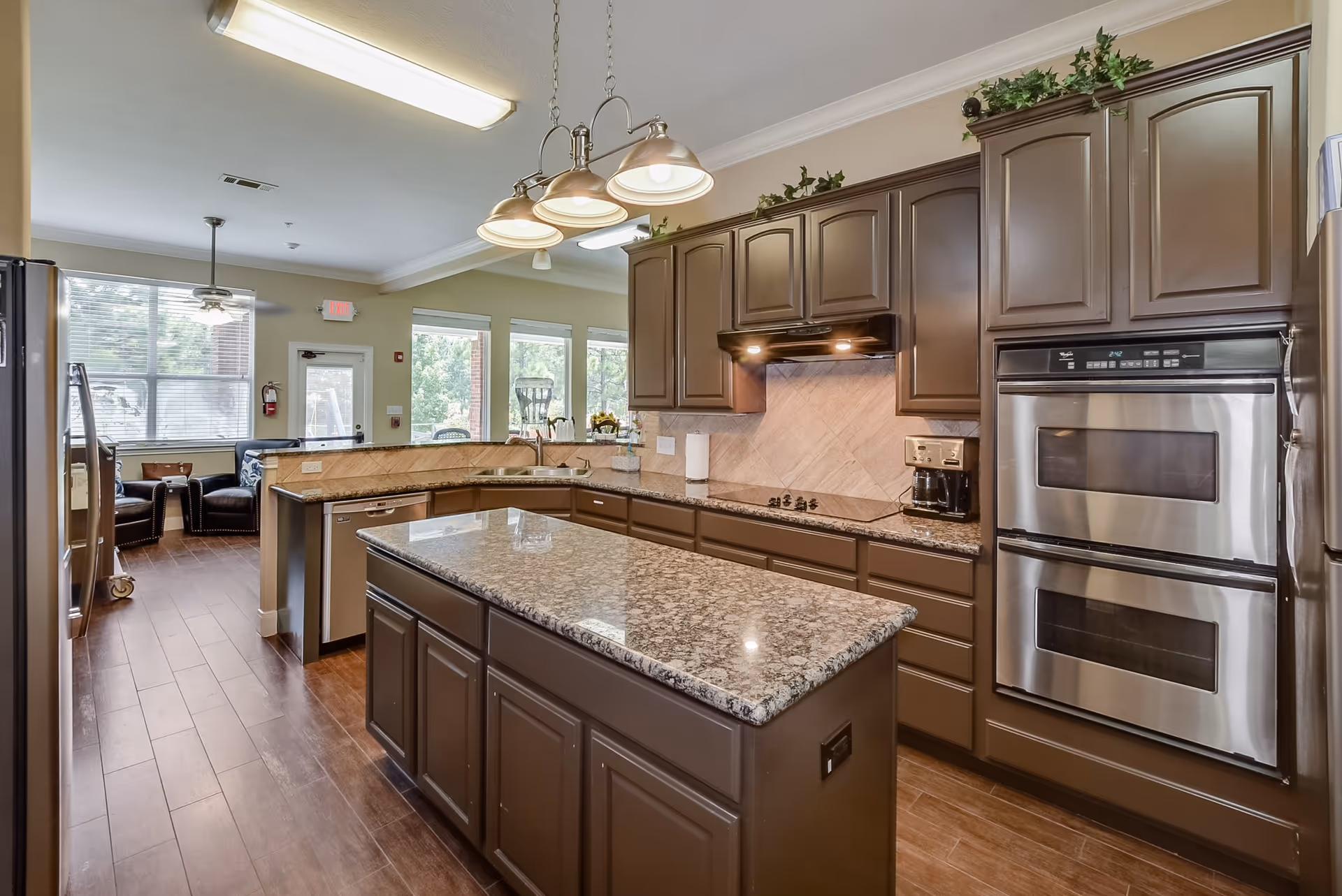 A modern kitchen with brown cabinets and granite countertops, featuring a central island, built-in double ovens, a stovetop with an overhead vent, a coffee maker, and a dishwasher. The kitchen opens into a seating area with large windows and two black armchairs.