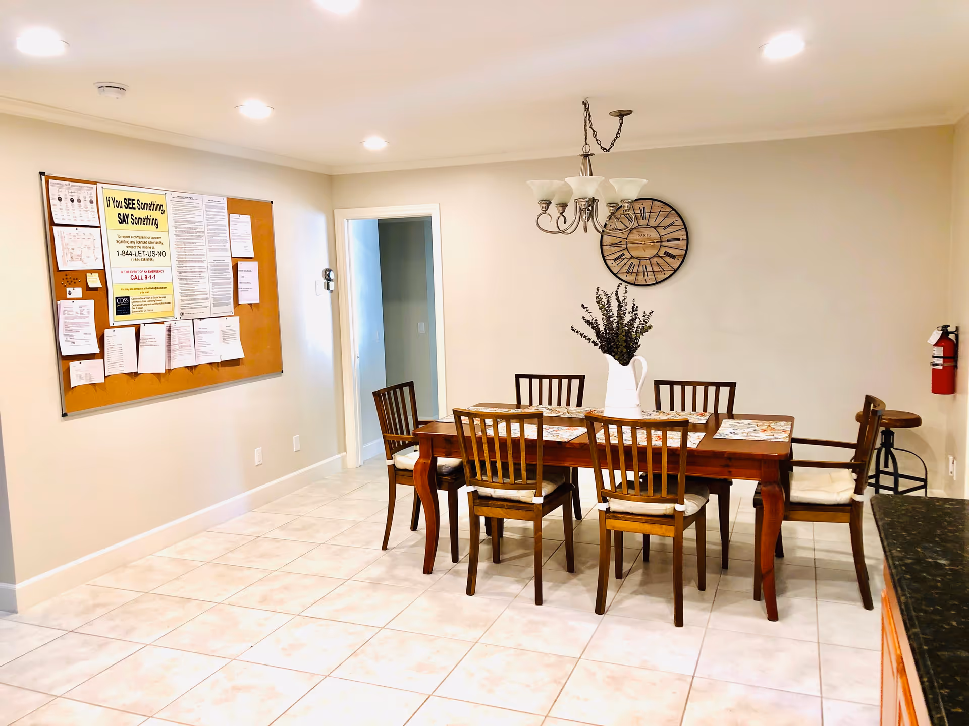 A dining area with a wooden table and six chairs, a white vase with flowers on the table, a large wall clock, a chandelier, a bulletin board with notices on the wall, and a fire extinguisher mounted on the wall.