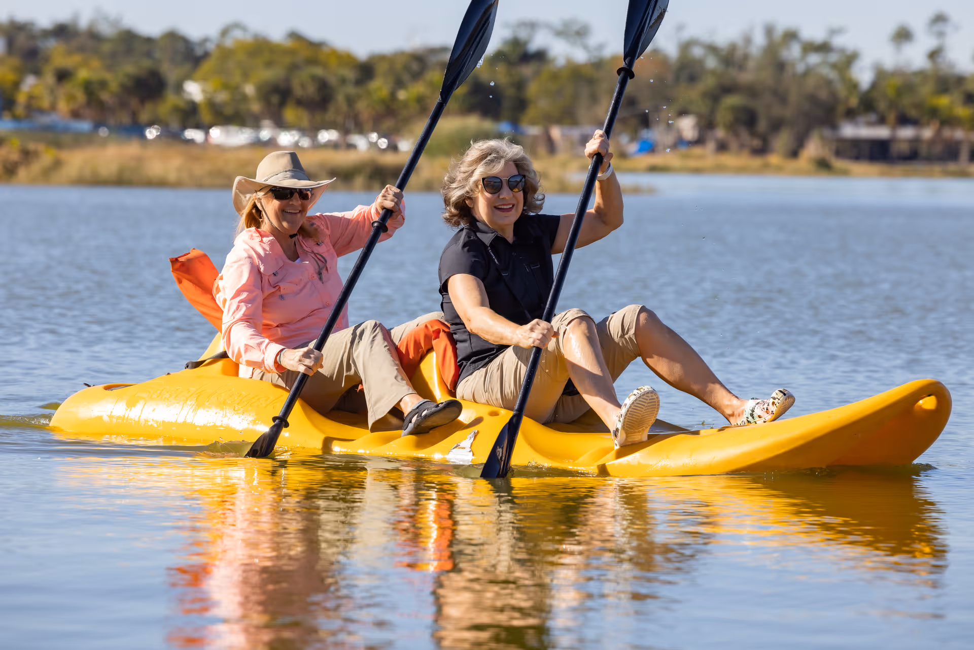 Two smiling older women paddling a yellow tandem kayak on a calm lake.