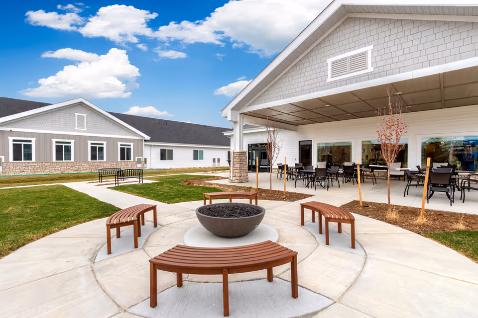 Outdoor patio area at Greeley Village featuring a circular fire pit surrounded by curved wooden benches, a covered seating area with tables and chairs, and a well-maintained lawn with pathways and small trees under a partly cloudy sky.