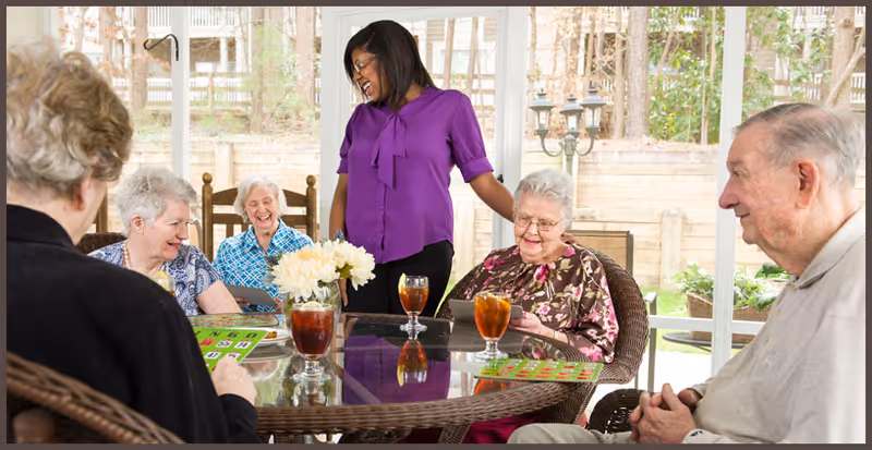 A group of elderly people sitting around a glass-top table playing bingo with a caregiver standing and smiling among them. The setting is a bright indoor area with large windows showing an outdoor garden.
