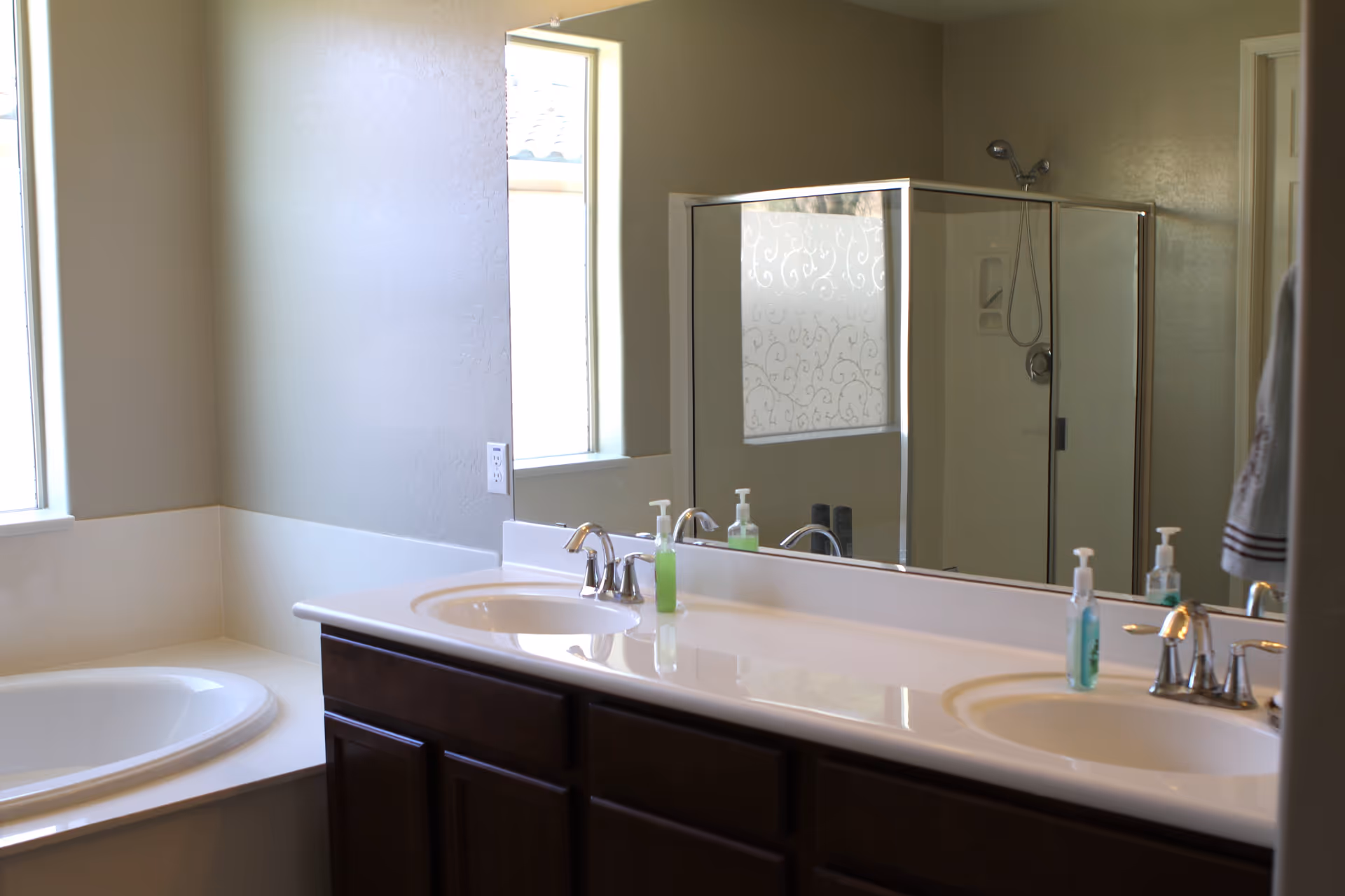 A bathroom with a double sink vanity featuring two faucets and soap dispensers, a large mirror above the sinks, a bathtub to the left, and a glass-enclosed shower reflected in the mirror. The walls are painted light beige, and there is a window letting in natural light.