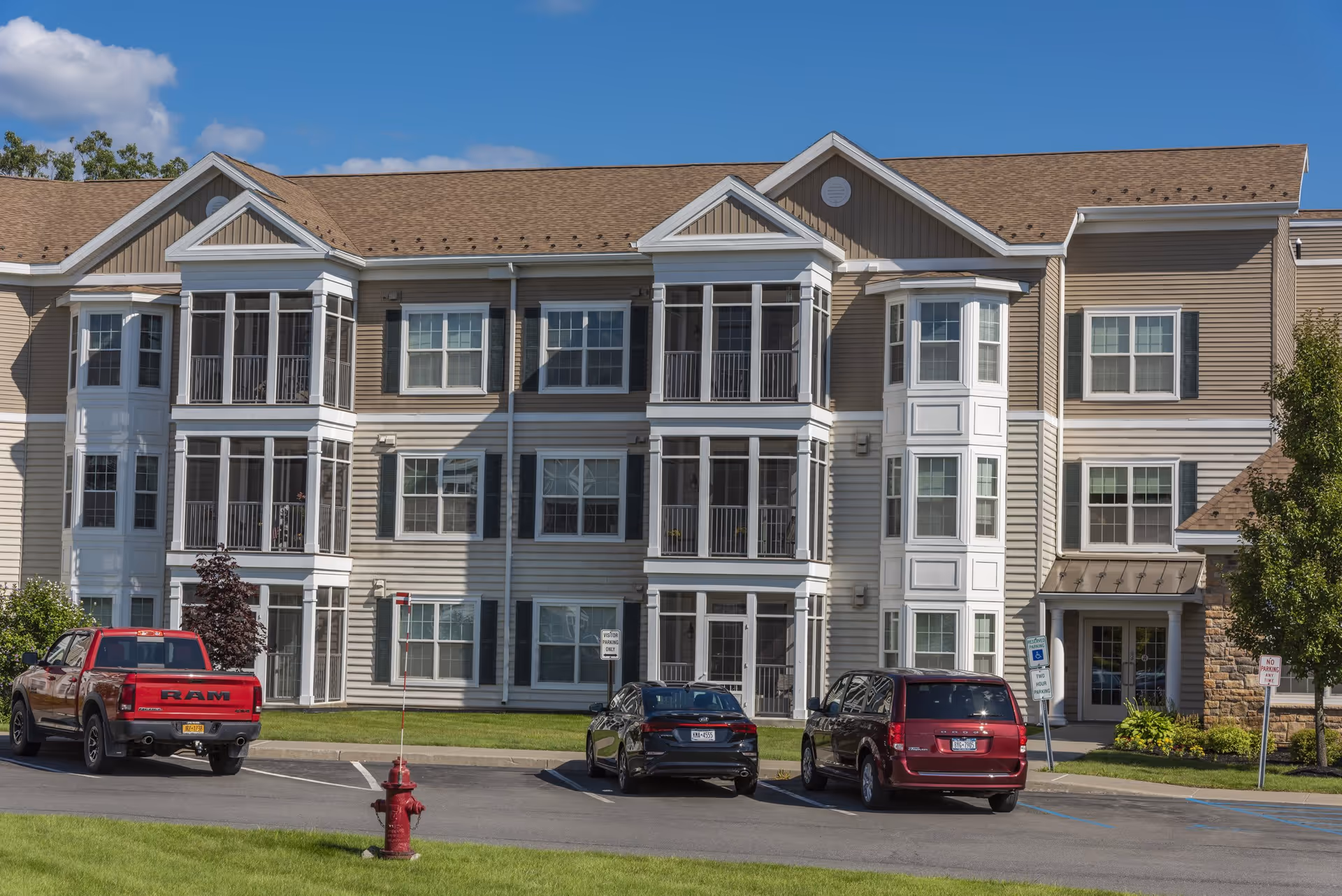 Exterior view of Coburg Village, a multi-story senior living facility with beige siding, white trim, and multiple large windows and screened balconies. There are three vehicles parked in front, a red RAM truck, a black sedan, and a maroon minivan. A red fire hydrant is visible on the grass near the parking lot.