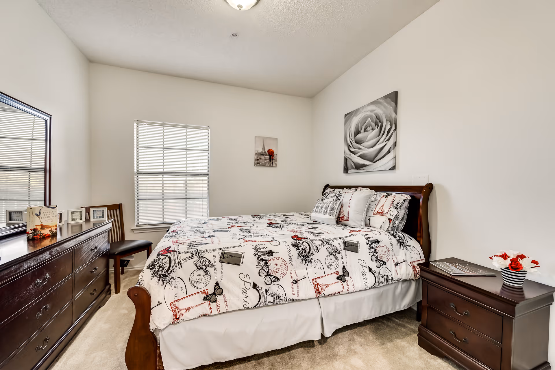 A bedroom with a wooden bed frame and a bed made with white bedding featuring black and red Paris-themed prints. There is a matching wooden dresser with a large mirror on the left side and a wooden nightstand with two drawers on the right side. The nightstand has a small vase with red and white flowers and a book on it. The room has beige carpet, white walls, a window with blinds, and two pieces of wall art: a large black and white rose above the bed and a smaller picture of a person with a red umbrella near the window.