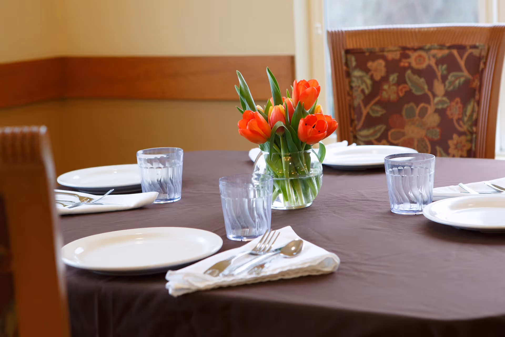 A round dining table set with white plates, silverware on white napkins, and clear drinking glasses. A small glass vase with bright orange tulips is placed at the center of the table. The background shows a window and a chair with a floral patterned cushion.
