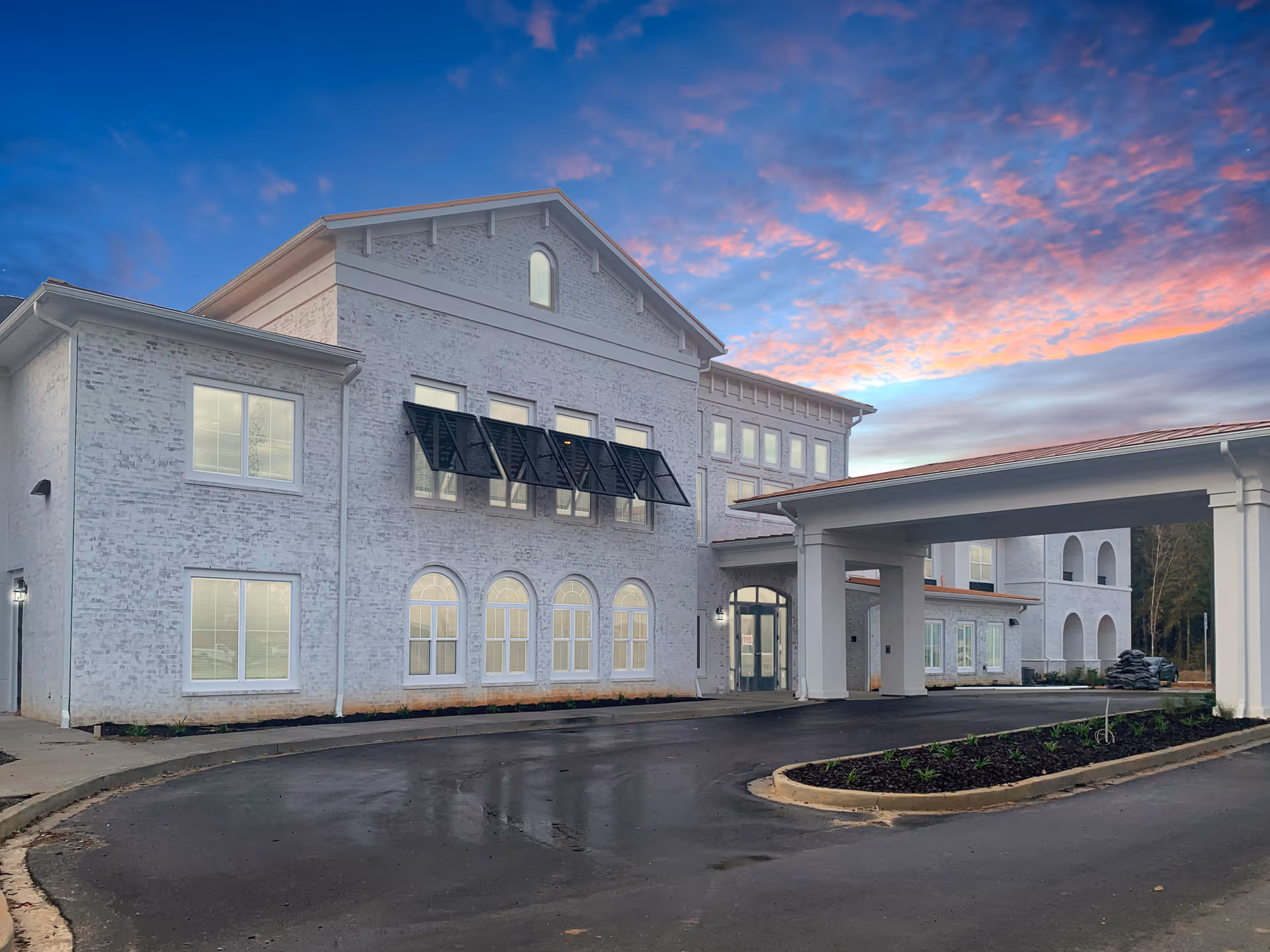 Exterior view of The Blake at Biloxi senior living facility during sunset, showing a large white brick building with multiple windows, a covered entrance driveway, and a landscaped area with plants.