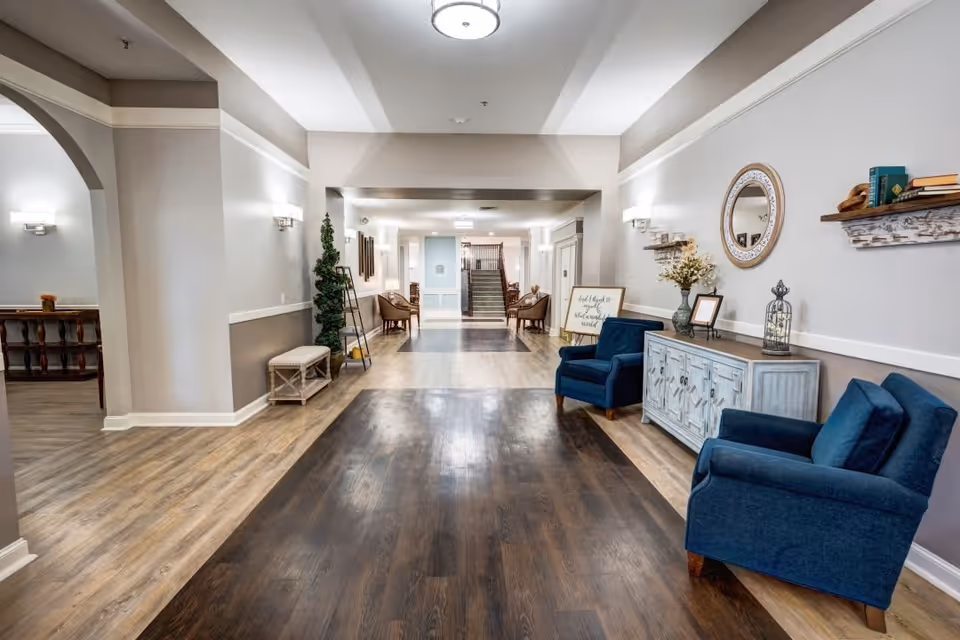 A spacious and well-lit hallway in a senior living facility with light-colored walls and wood flooring. The hallway features blue upholstered armchairs, a decorative sideboard with a round mirror above it, and shelves with books and decor items. There are additional seating areas with chairs and small tables further down the hallway, and a staircase is visible at the end.