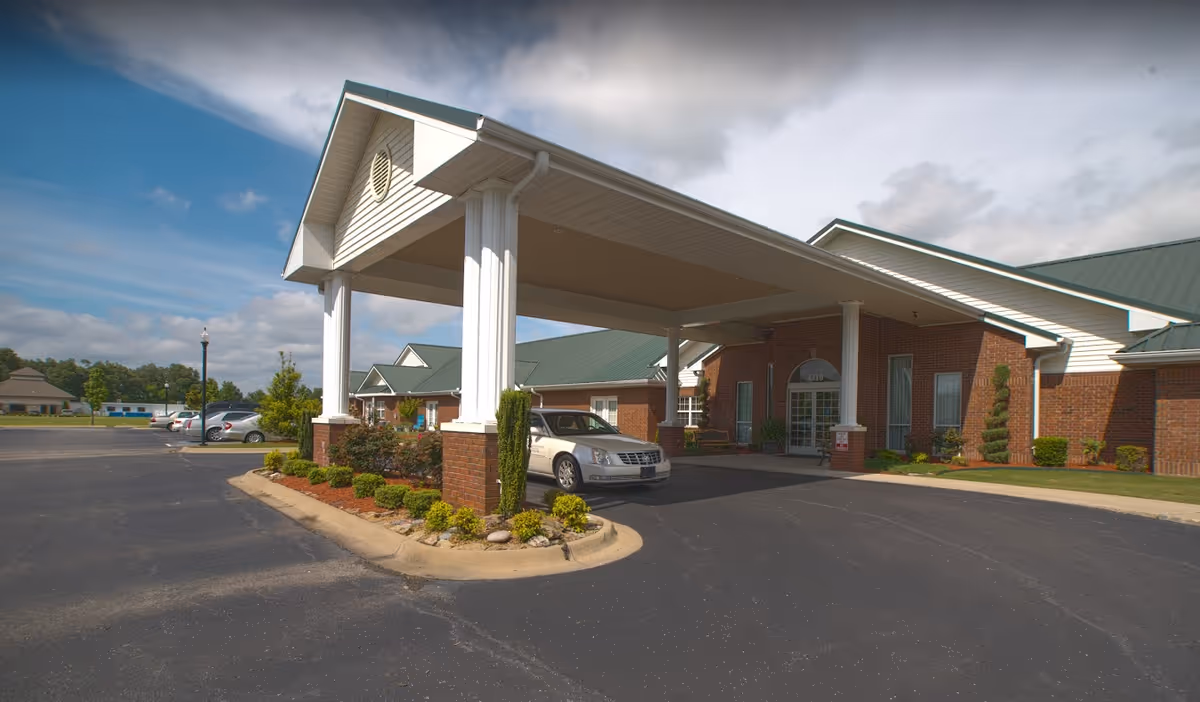 Brick senior living building front with a covered drop-off porte-cochère, landscaped island, and a car parked under the canopy.