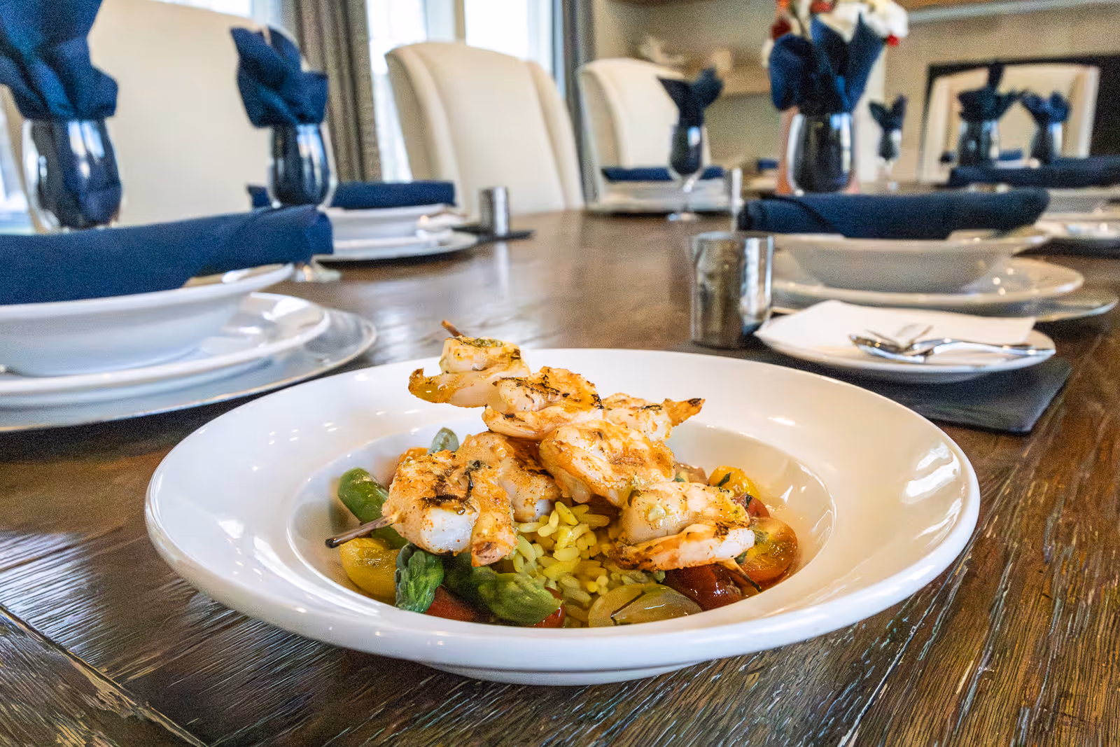 Grilled shrimp skewers served over rice and vegetables in a white bowl on a set dining table with plates and blue napkins.
