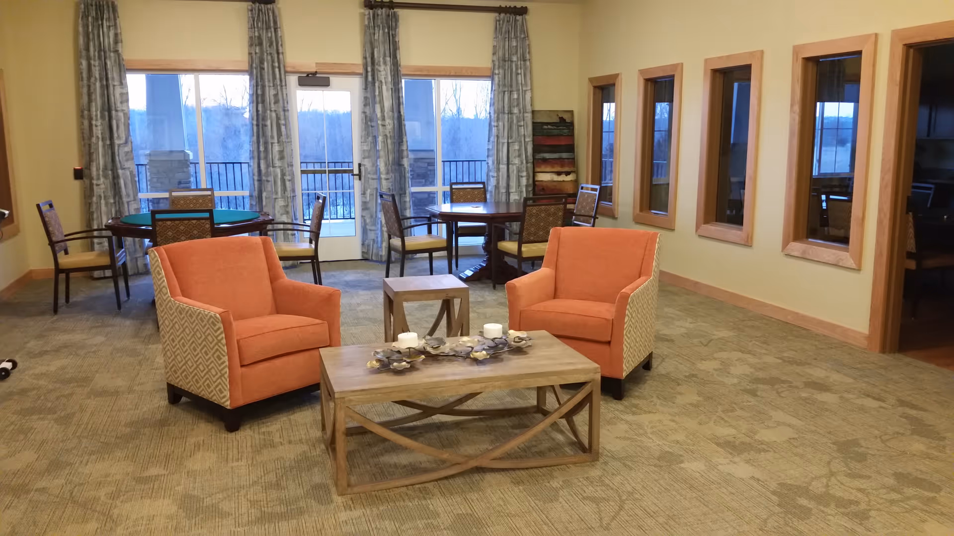 A cozy sitting area in a senior living facility with two orange armchairs facing a wooden coffee table decorated with candles and a centerpiece. Behind the chairs, there are two round tables with chairs near large windows and glass doors with curtains, allowing natural light to enter. The walls have three rectangular windows with wooden frames, and the room has a carpeted floor.