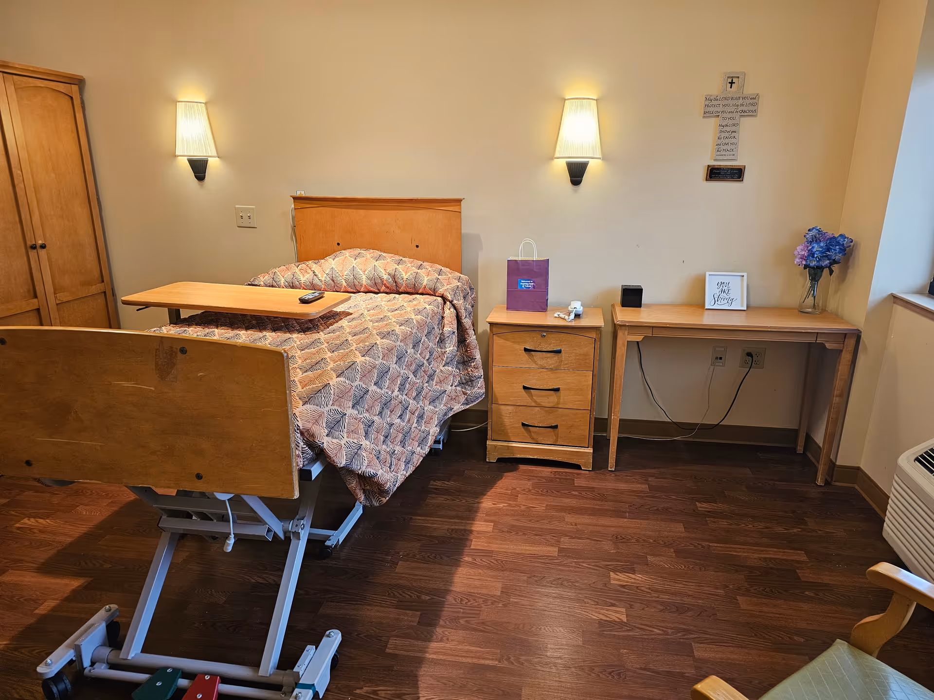 A senior care facility bedroom with a hospital-style adjustable bed covered with a patterned quilt. There is a wooden nightstand with three drawers next to the bed, a wooden desk with a vase of flowers and a framed quote, and two wall-mounted lamps above the bed. The room has wood flooring and light-colored walls.