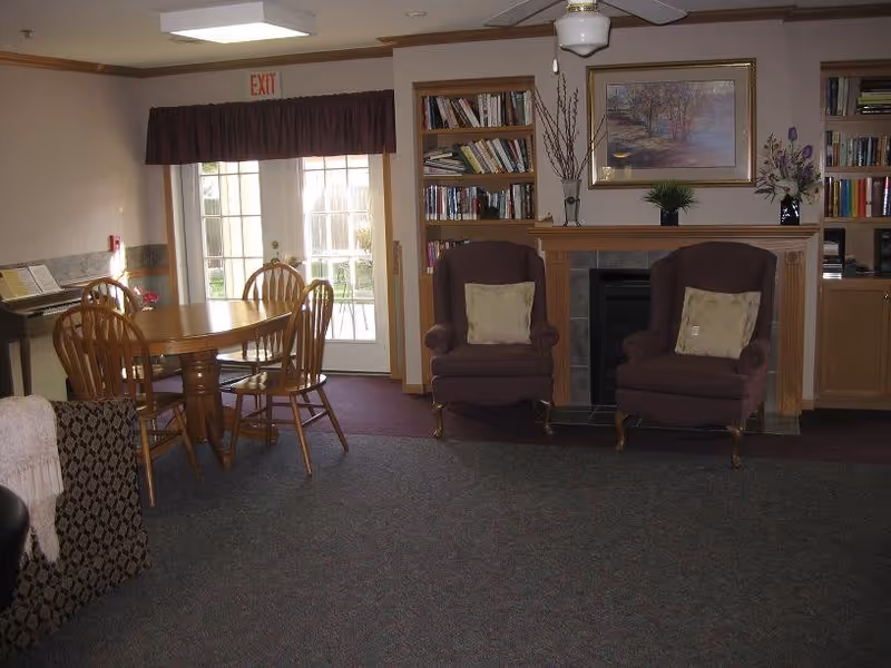 A cozy living area featuring two upholstered armchairs with cushions in front of a fireplace. Above the fireplace is a framed painting and decorative plants. To the left, there is a wooden dining table with four chairs near glass double doors with an exit sign above. Bookshelves filled with books flank the fireplace, and a piano is partially visible on the far left.