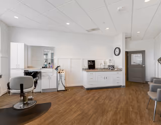 Bright interior salon area with a barber chair, wash station, cabinetry, and seating in a clean common room.