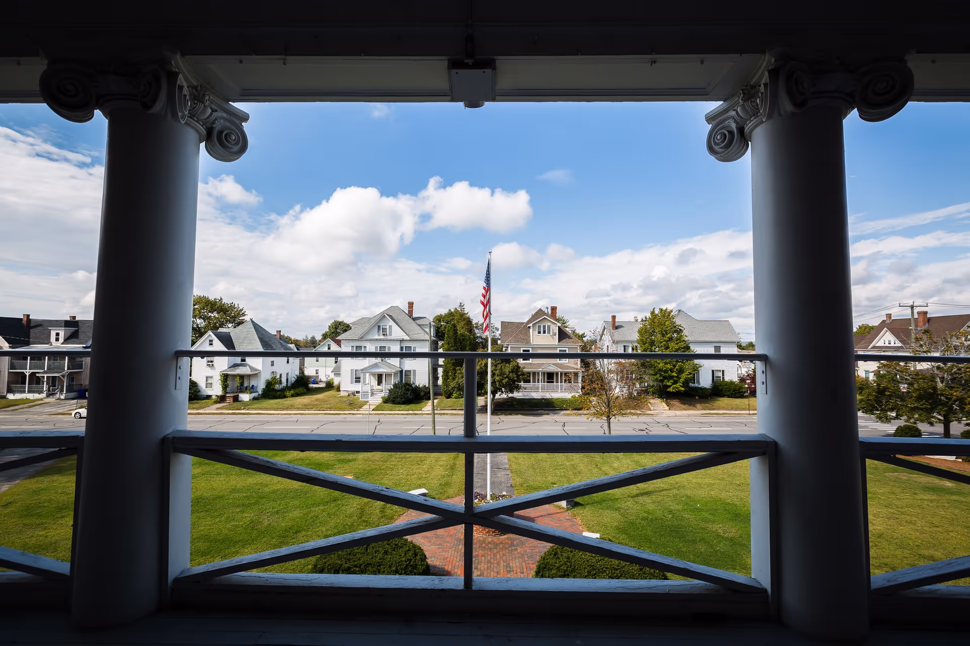 View from a porch or balcony framed by two large white columns and a wooden railing, overlooking a green lawn with a brick pathway and an American flagpole in the center. Across the street are several residential houses under a partly cloudy blue sky.