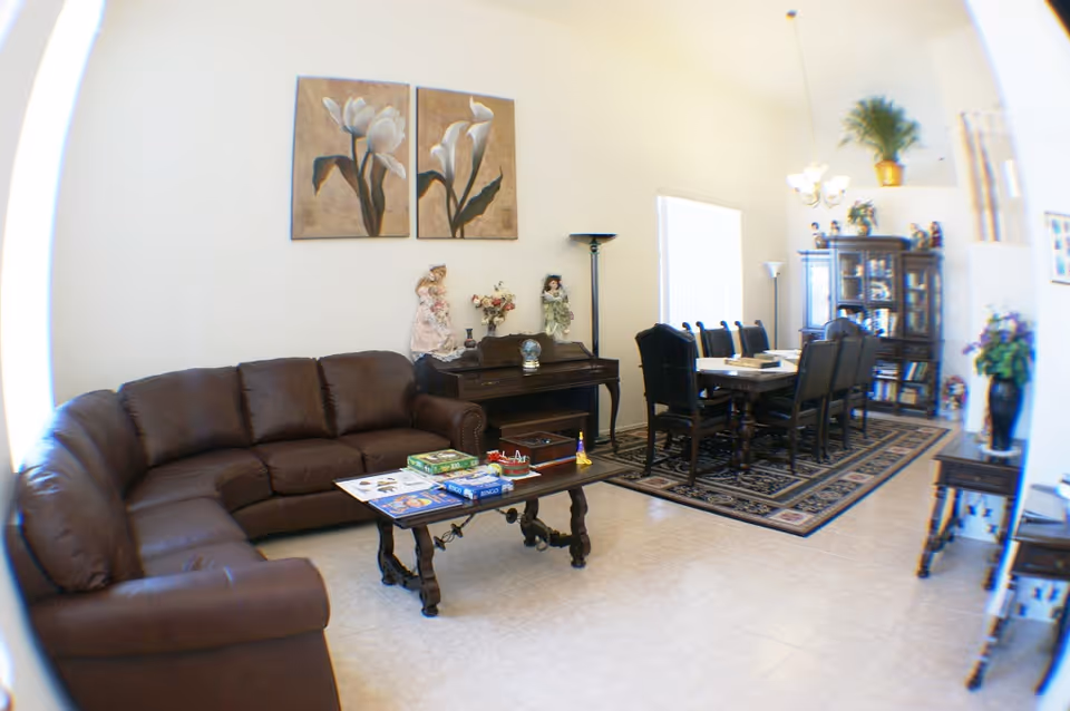 A spacious living and dining area with a brown leather sectional sofa, a wooden coffee table with board games on it, a piano with decorative dolls and flowers, and a dining table with eight chairs on a patterned rug. The room has light-colored tiled floors, high ceilings, and wall art featuring flowers.