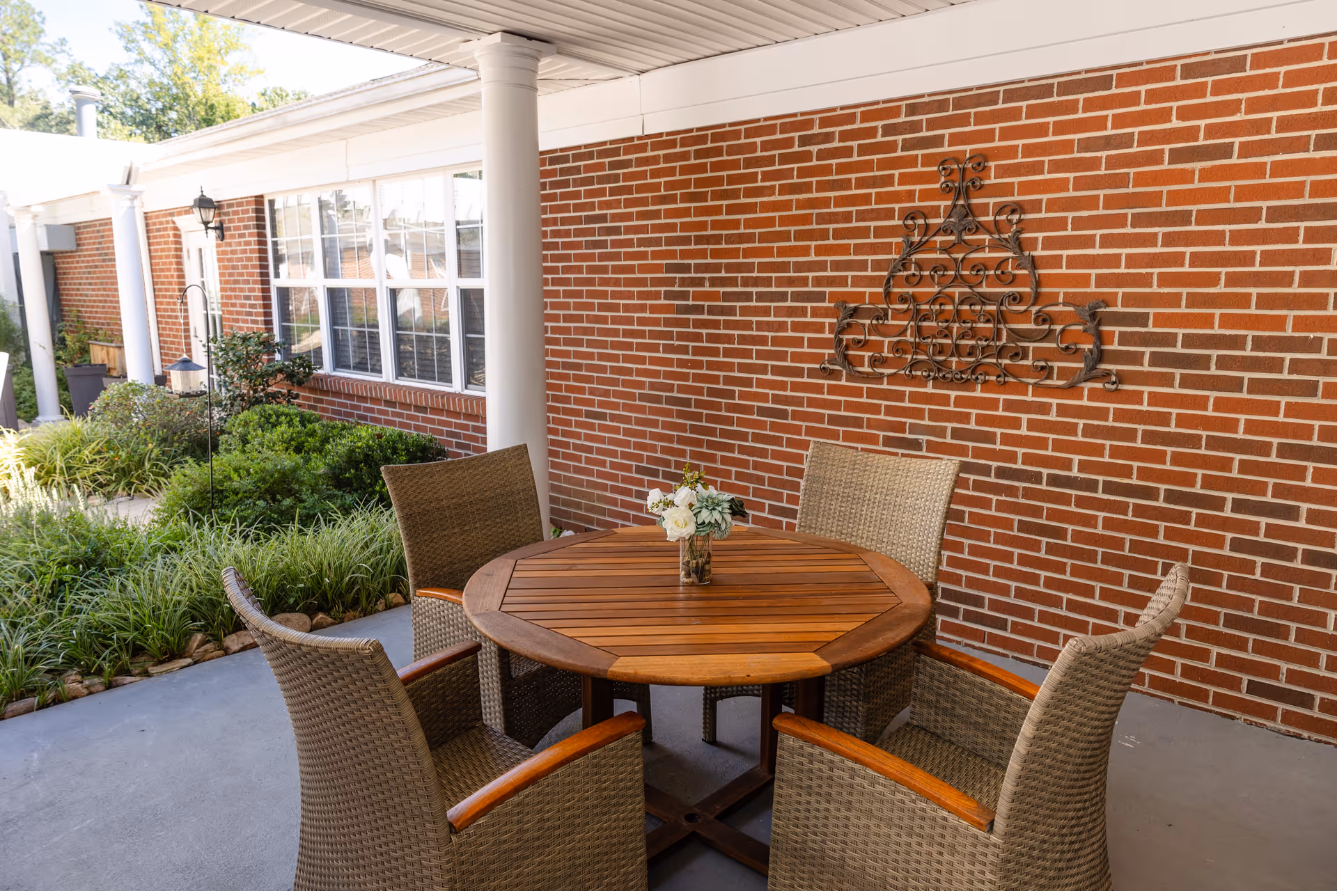 Covered outdoor patio area with a round wooden table surrounded by four wicker chairs. A small vase with flowers is placed on the table. The patio is adjacent to a brick wall with decorative metal wall art and white columns supporting the roof. Green shrubs and plants are visible along the edge of the patio.