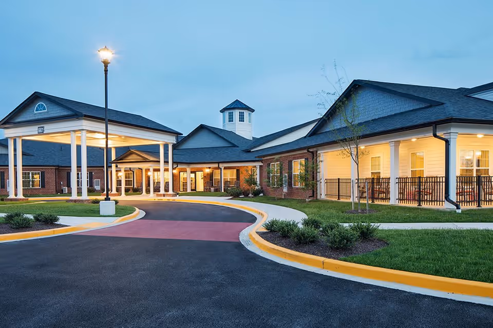 Exterior view of Spring Arbor of Salisbury senior living facility at dusk, showing a well-lit entrance with a covered driveway, manicured landscaping, and a combination of brick and siding on the building.