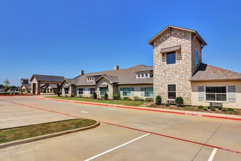 Front exterior of a single-story senior living facility with a stone tower feature and an empty parking lot under a clear blue sky.