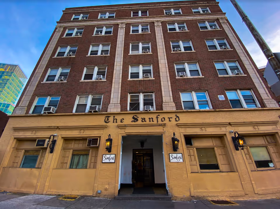 Exterior view of a multi-story brick building named The Sanford with a yellow lower facade and multiple windows, some with air conditioning units. The entrance is centered with two black lantern-style lights on either side and signs that read 'Sanford'.
