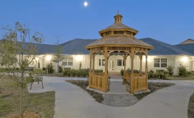 Outdoor view of a senior living facility courtyard at dusk featuring a wooden gazebo in the center with benches inside. The building surrounds the courtyard with windows and exterior lights illuminating the area. There are paved walkways and some landscaping with small trees and bushes.