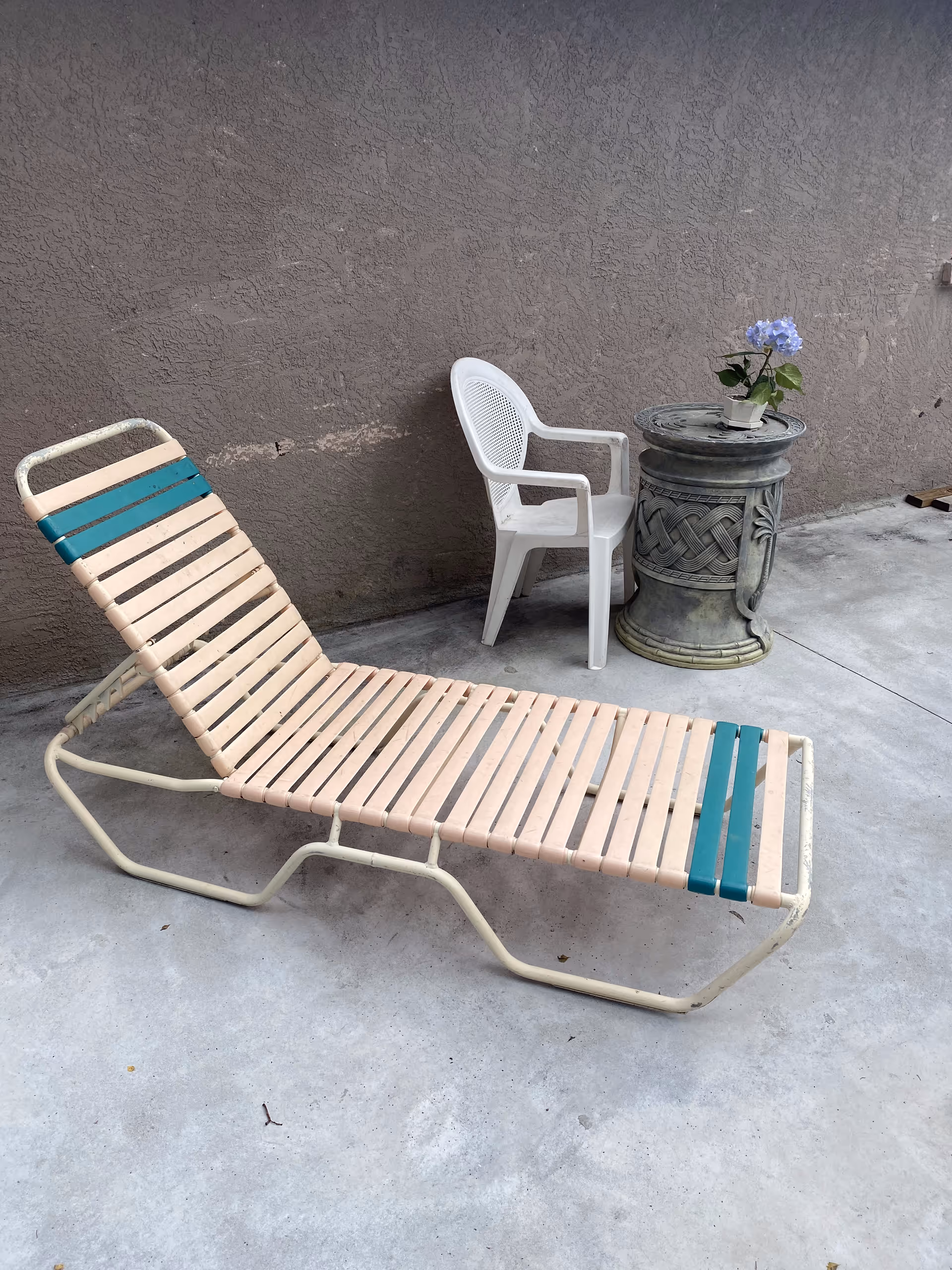 Outdoor patio area with a beige and teal lounge chair, a white plastic chair, and a decorative round stone table with a small potted plant with purple flowers on top, against a textured brown wall.
