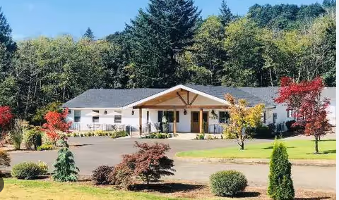 Single-story white building with a dark roof surrounded by a paved driveway and landscaped greenery including small trees and bushes, set against a backdrop of tall trees and a clear sky.