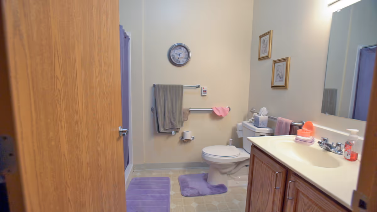 A bathroom with a wooden door partially open, showing a toilet with a purple bath mat in front, a white sink with a wooden cabinet, a large mirror above the sink, a wall clock, and two framed pictures on the wall. There are towels hanging on metal bars and a purple shower curtain visible in the background.