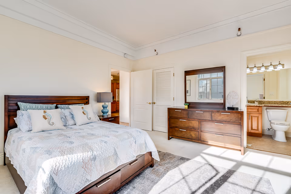 A bright bedroom with a large bed featuring a wooden headboard and sea-themed bedding. There is a wooden dresser with a mirror on top, a bedside table with a blue lamp, and an open door leading to a bathroom with a toilet and vanity. Sunlight streams through a window, casting shadows on the carpeted floor.