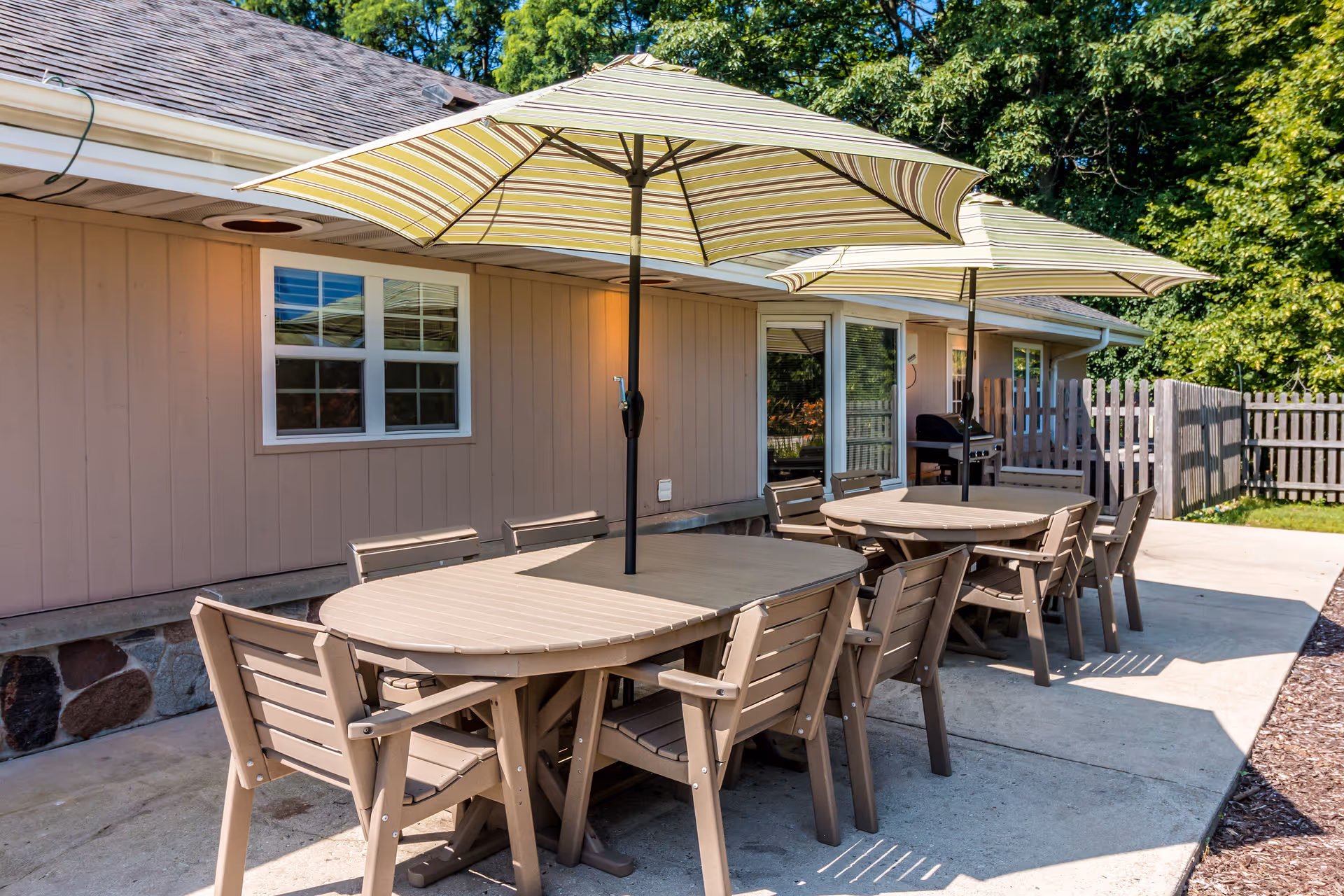 Outdoor patio area with two beige tables, each surrounded by six matching chairs. Each table has a large striped umbrella providing shade. The patio is adjacent to a beige building with windows and a door, and there is a wooden fence and greenery in the background.
