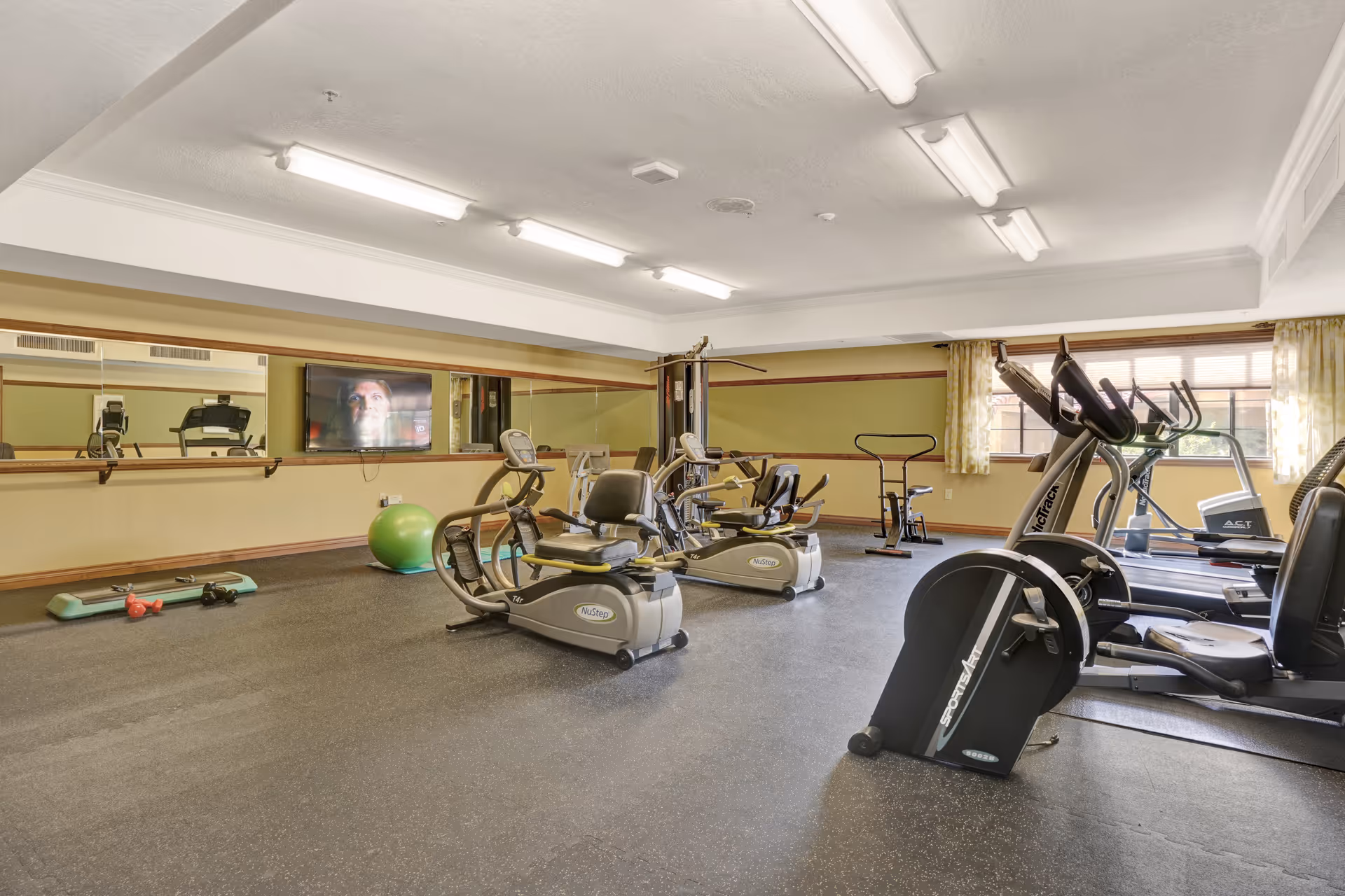 A fitness room with various exercise equipment including recumbent bikes, treadmills, a multi-station gym machine, a green exercise ball, dumbbells, and a step platform. The room has a large mirror on one wall, a wall-mounted TV, and windows with yellow patterned curtains letting in natural light.