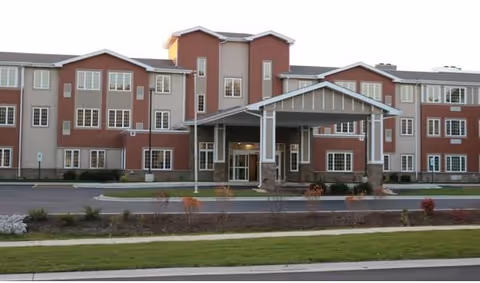 Front exterior view of Lacey Creek Supportive Living facility, a large three-story building with a covered entrance, multiple windows, and a landscaped area with grass and small shrubs in front.