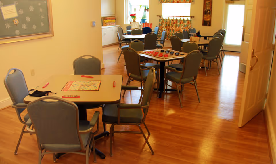 A well-lit room with several tables and chairs arranged for group activities. One table has a Scrabble board game set up, and another table has a checkers game. The room has wooden flooring, a bulletin board on the wall, and windows with colorful curtains letting in natural light.