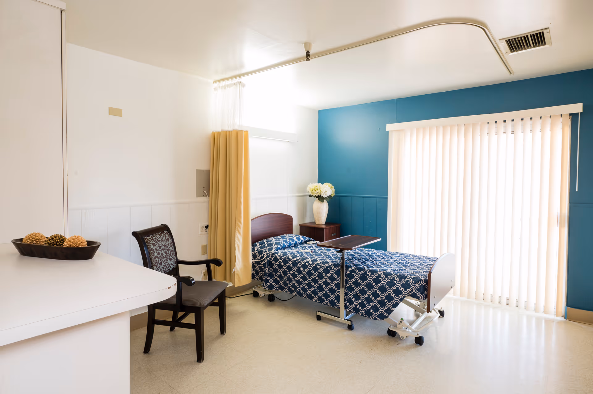 A bright and clean patient room with a hospital bed covered in a blue patterned blanket, a wooden chair with patterned upholstery, a small bedside table with a vase of white flowers, and vertical blinds covering a large window or sliding door. There is a yellow privacy curtain partially drawn and a white countertop with decorative pine cones on the left side.
