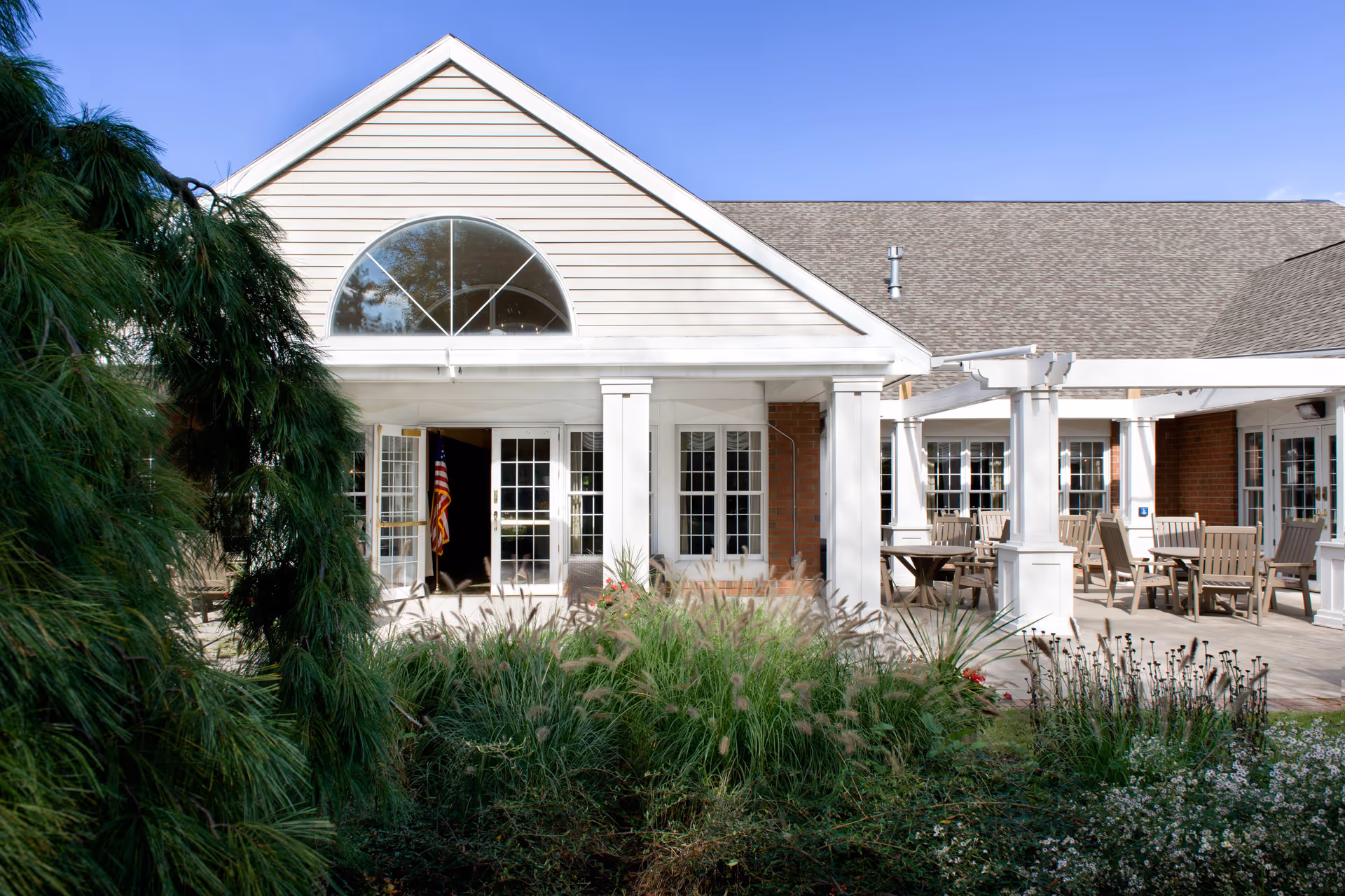 Exterior view of a senior living facility building with beige siding and a large arched window above double glass doors. There is a covered patio area with wooden tables and chairs, surrounded by greenery and plants in the foreground. The sky is clear and blue.