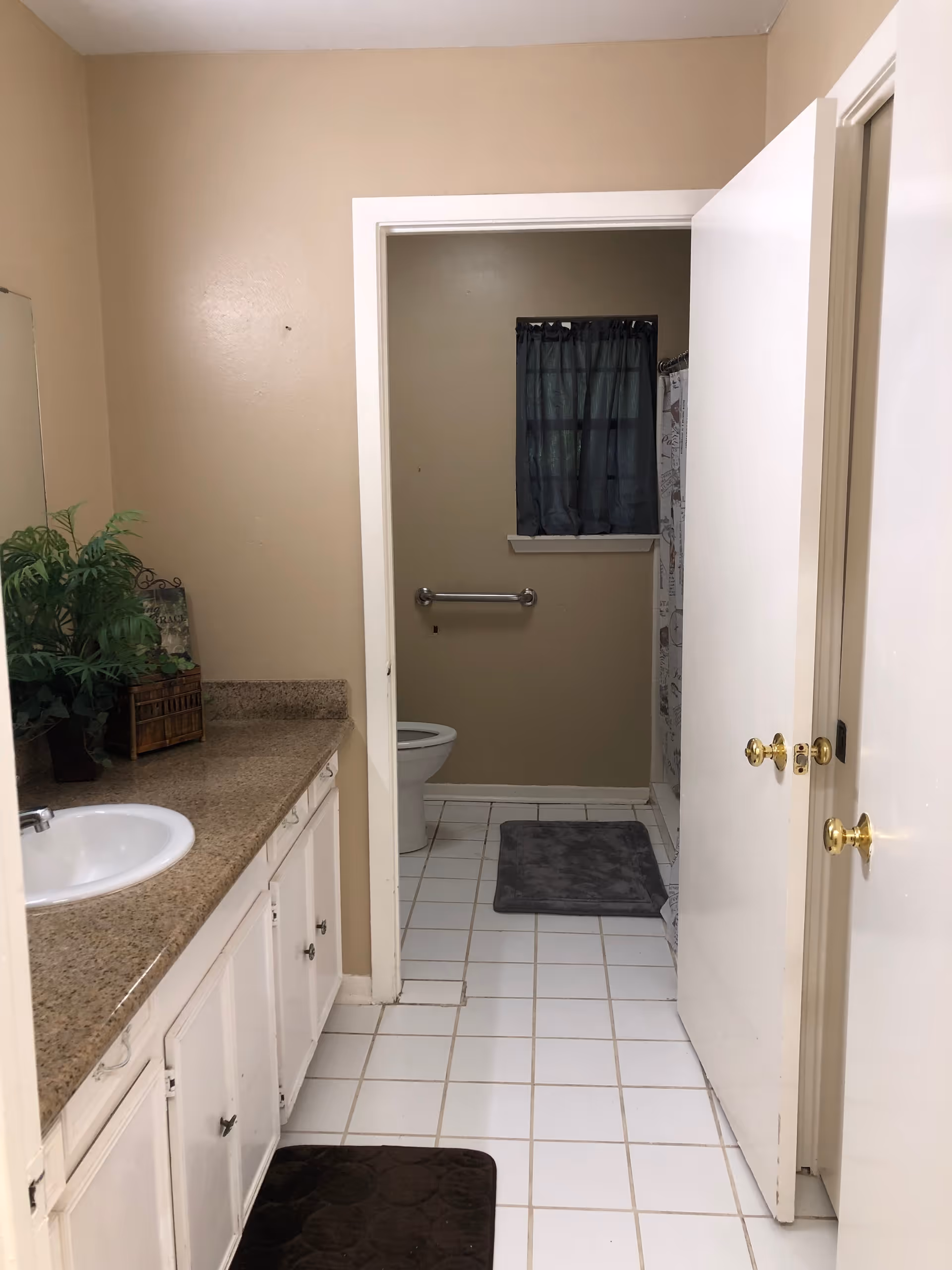 Bathroom interior showing a vanity with sink on the left, tiled floor, and an open door leading to a toilet and shower area with a grab bar.