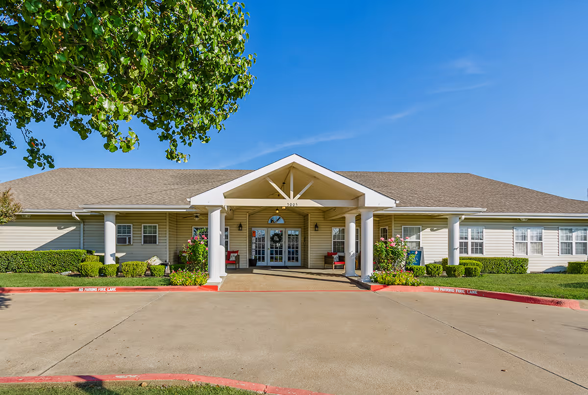 Front exterior view of a single-story building with a covered entrance supported by white columns, surrounded by green shrubs and flowers under a clear blue sky.