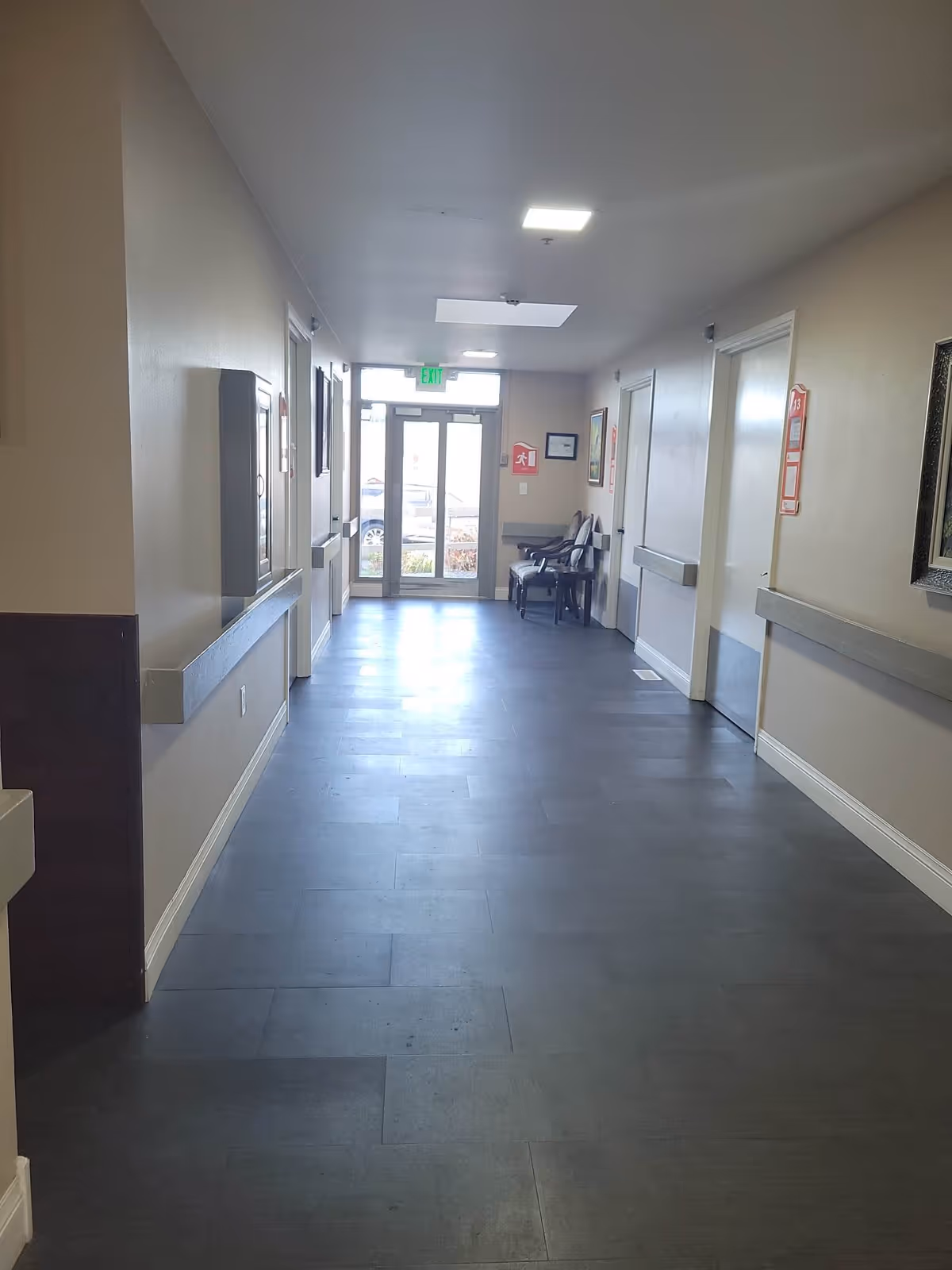 A clean, well-lit hallway in a senior living facility with beige walls, dark flooring, and several closed doors on both sides. There are chairs and framed pictures along the right wall near the glass exit doors at the end of the hallway.