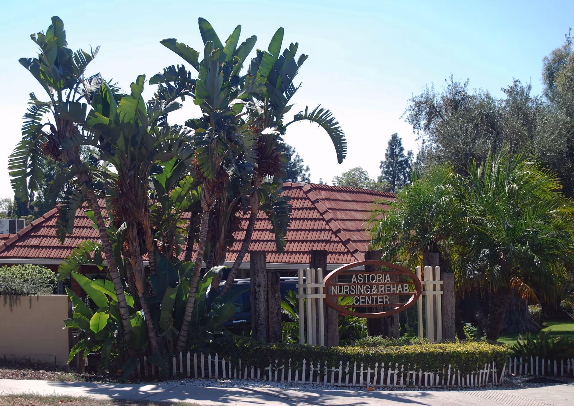 Exterior view of Astoria Nursing and Rehabilitation Center with tropical plants and trees in front of a building with a red tiled roof and a wooden sign displaying the facility name.