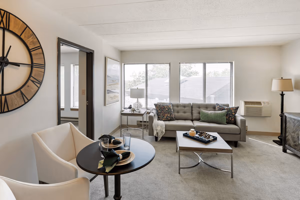 A bright living room in a senior living facility with a gray tufted sofa adorned with decorative pillows, a modern coffee table with a tray and candle, a small round dining table set for two with plates and glasses, a large wall clock, and two lamps providing ambient lighting. Large windows allow natural light to fill the space.