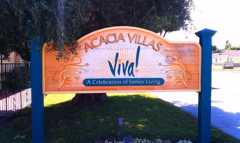 Wooden sign for Acacia Villas with the text 'Viva! A Celebration of Senior Living' mounted on two blue posts, with trees and a building in the background.
