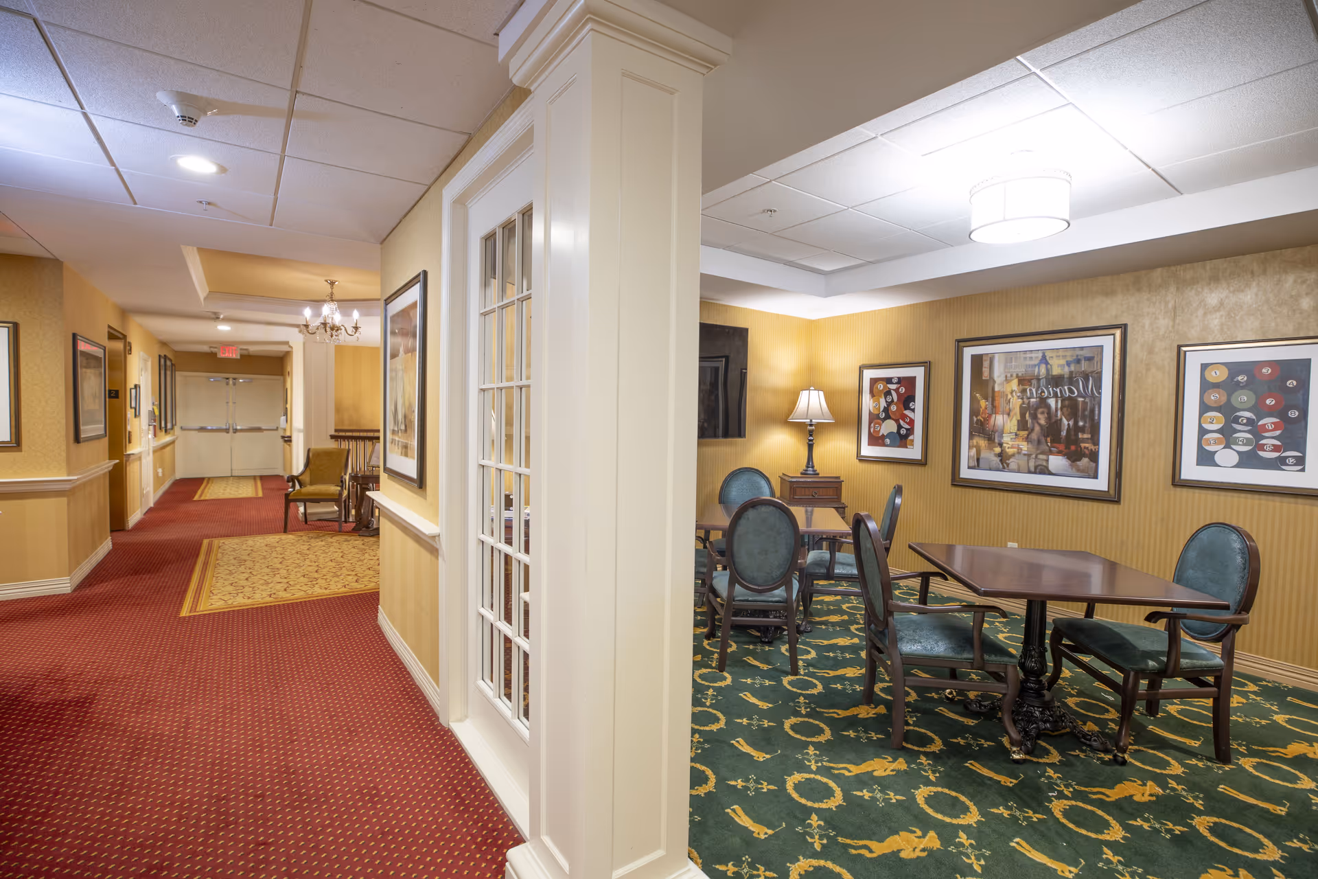 Interior view of a senior living facility showing a hallway with red carpet and yellow walls on the left, and a small sitting area with green carpet, wooden tables, and green upholstered chairs on the right. The sitting area has framed artwork on the walls and a table lamp on a small wooden side table.