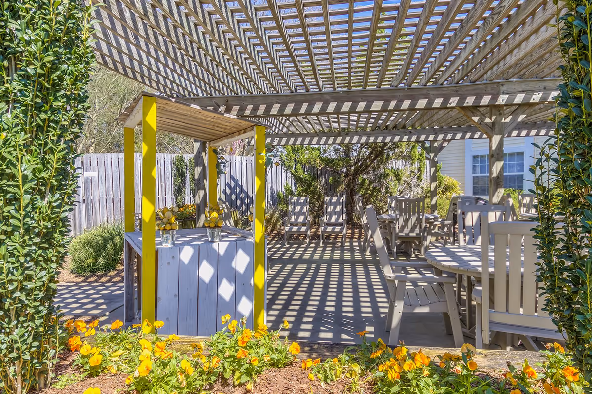 Shaded outdoor patio with a wooden pergola, tables and chairs, yellow-accented stand and flower beds.