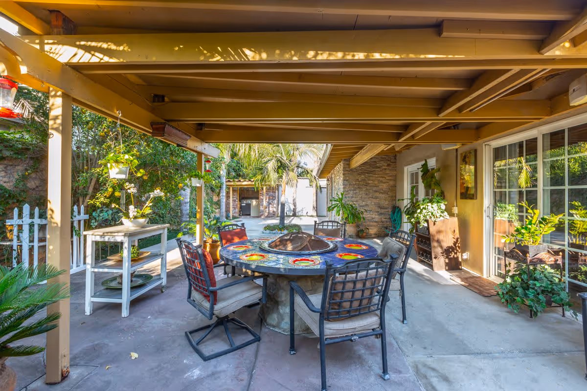 Covered outdoor patio area with a round table featuring colorful plates and a fire pit in the center, surrounded by six cushioned chairs. The patio is decorated with various potted plants and hanging planters, with a white picket fence and greenery in the background. Sliding glass doors lead inside the building.