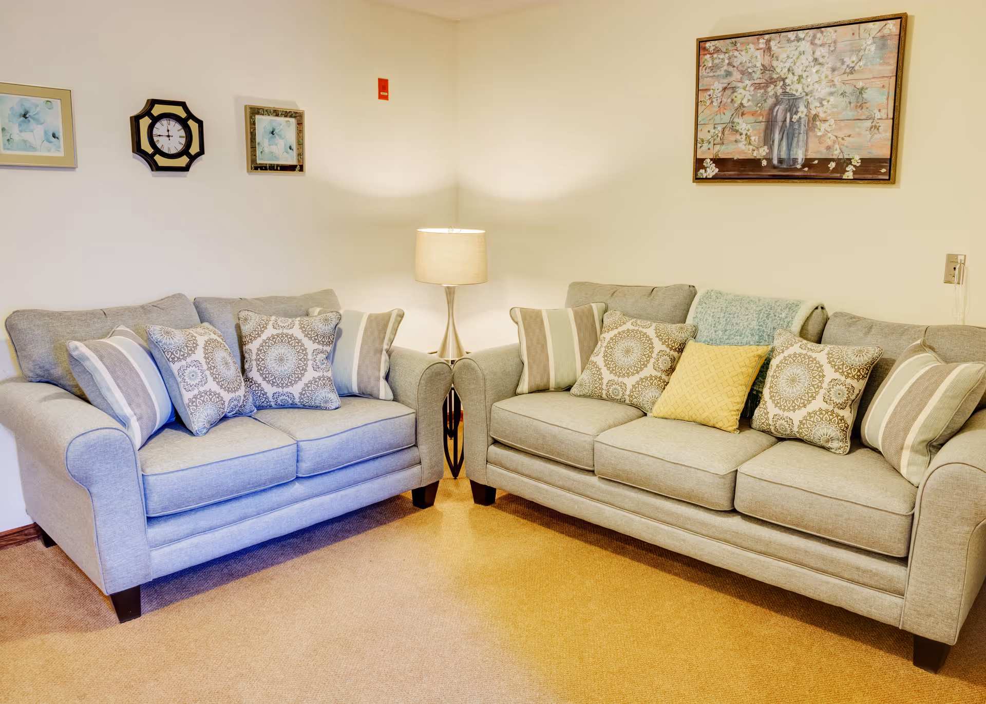Two gray sofas with patterned throw pillows arranged around a lamp in a cozy living room with wall art.