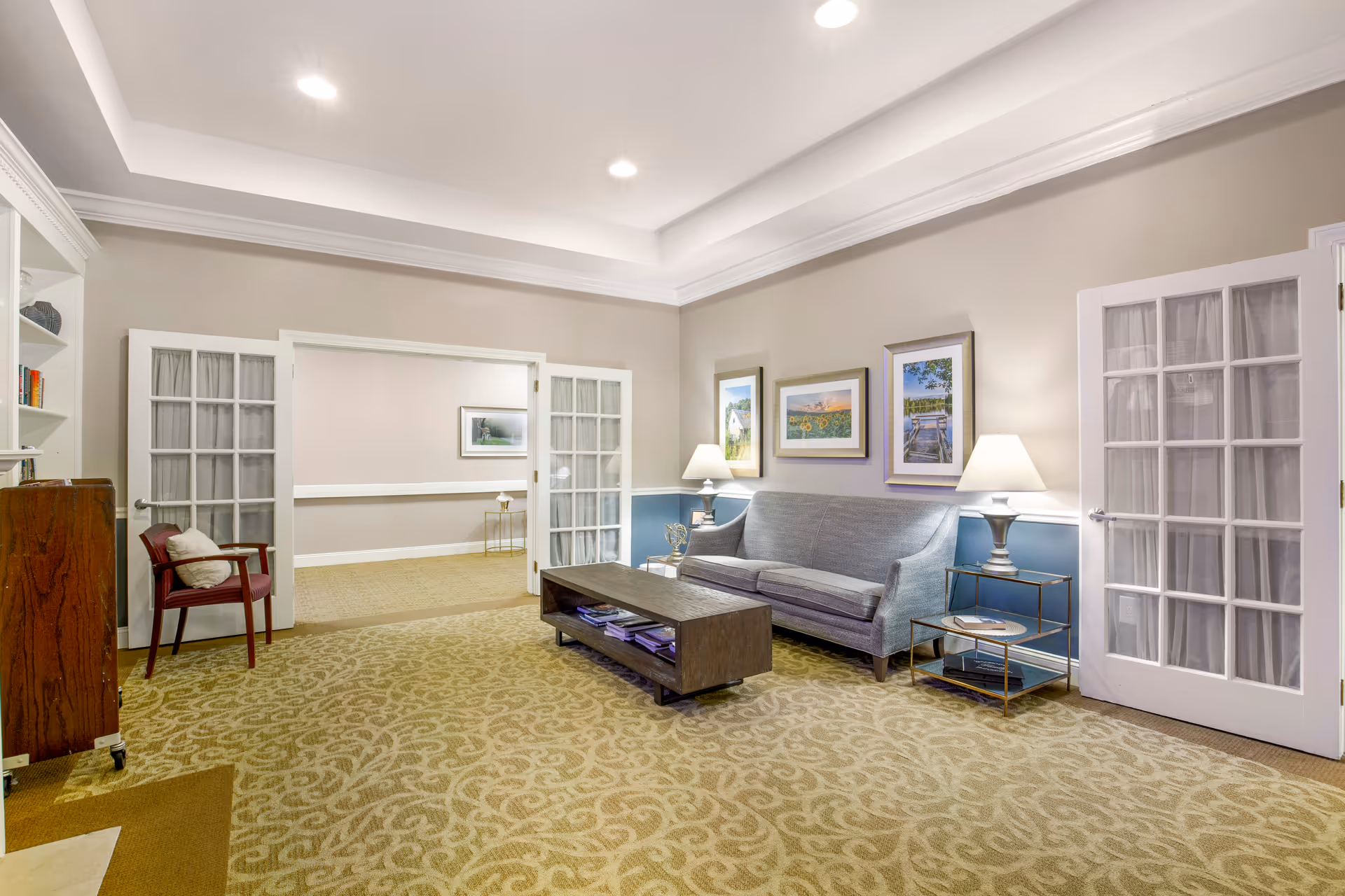 A well-lit living room area with a gray sofa, two side tables with lamps, a wooden coffee table with magazines underneath, and framed artwork on the wall. The room has beige patterned carpet and light-colored walls with white trim. French doors with glass panes lead to an adjacent room with similar decor.