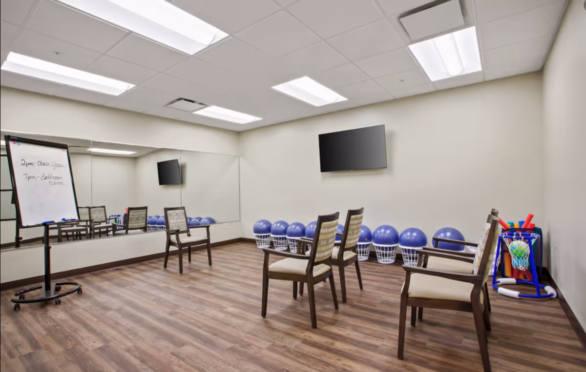 Activity/exercise room with chairs facing a wall-mounted TV, baskets of exercise balls, a mirrored wall, and a whiteboard.